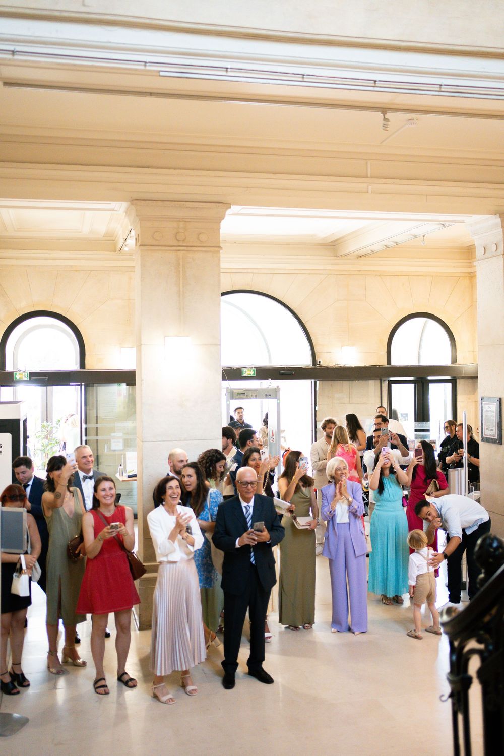 Wedding guests in formal attire gather in an elegant hall with arched windows, celebrating with champagne.