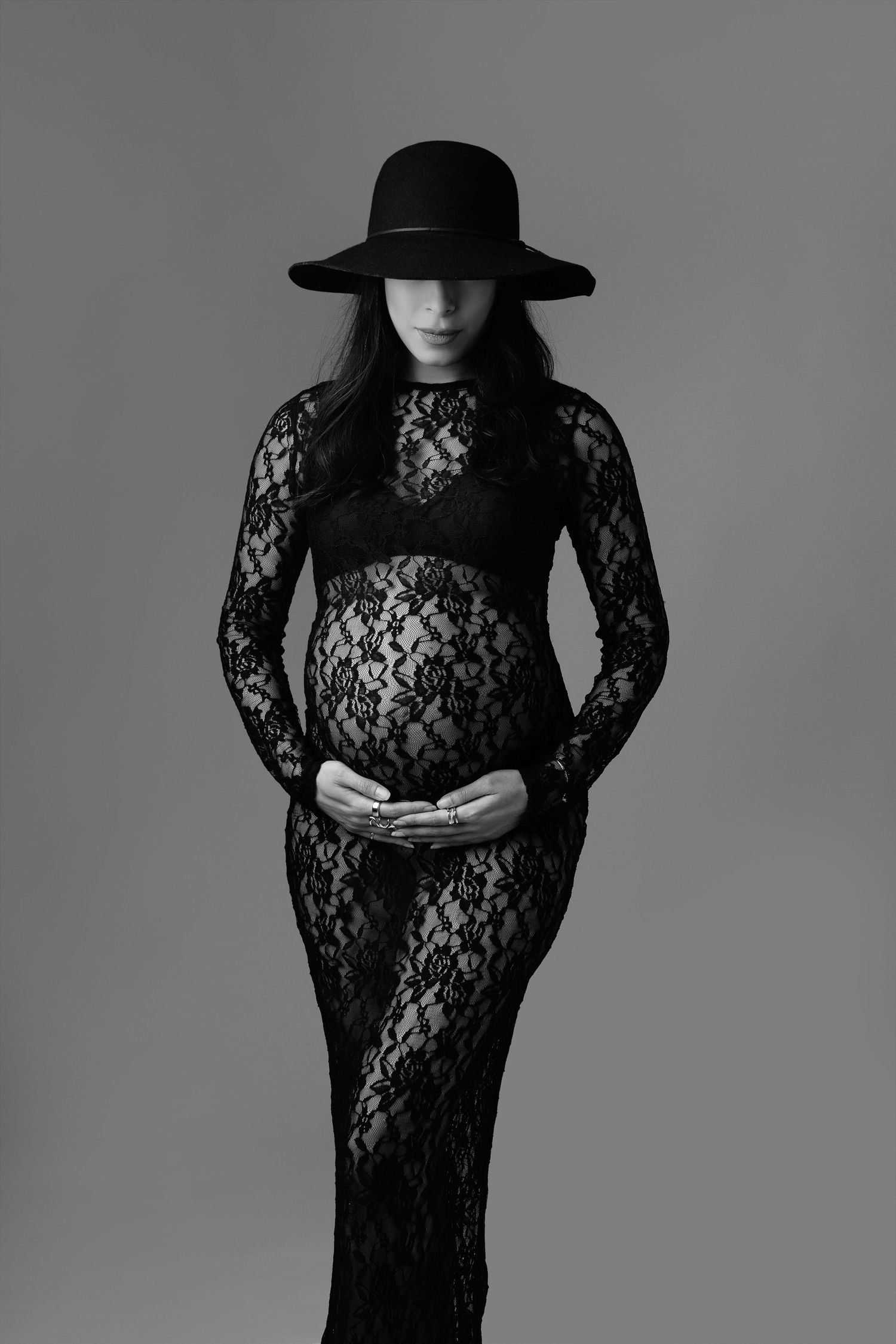 Pregnant woman in black lace dress and wide-brim hat cradling baby bump, elegant maternity portrait in black and white.