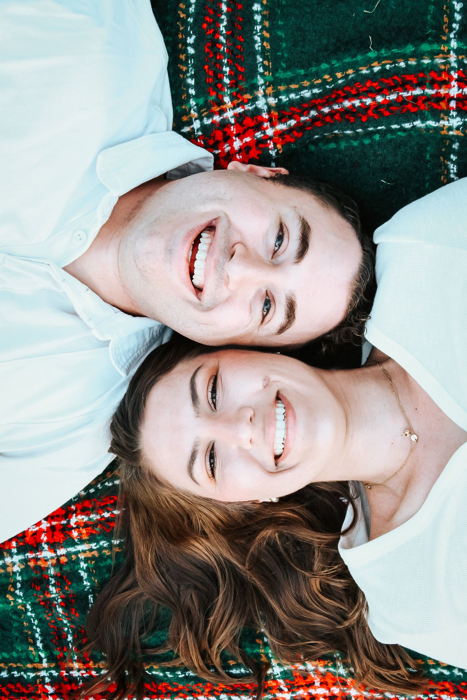 Overhead view of smiling couple lying on red and green plaid blanket looking up at camera.