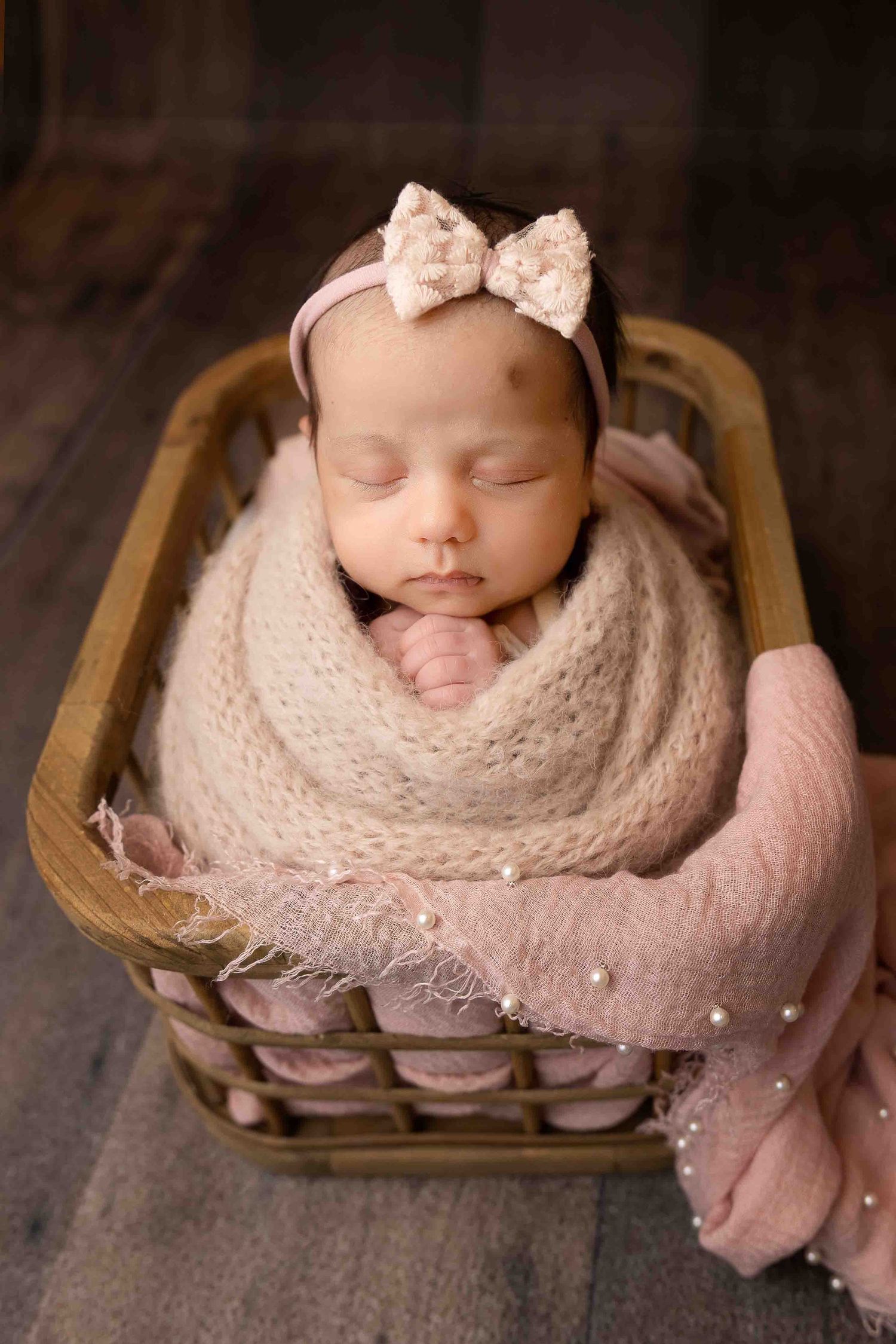Sleeping newborn baby girl wrapped in pink knit swaddle with bow headband, nestled in a wicker basket.