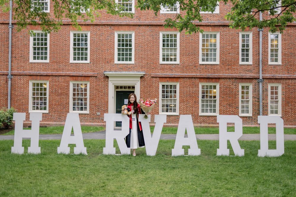 Harvard graduate in cap and gown poses proudly in front of large white HARVARD letters on campus lawn.