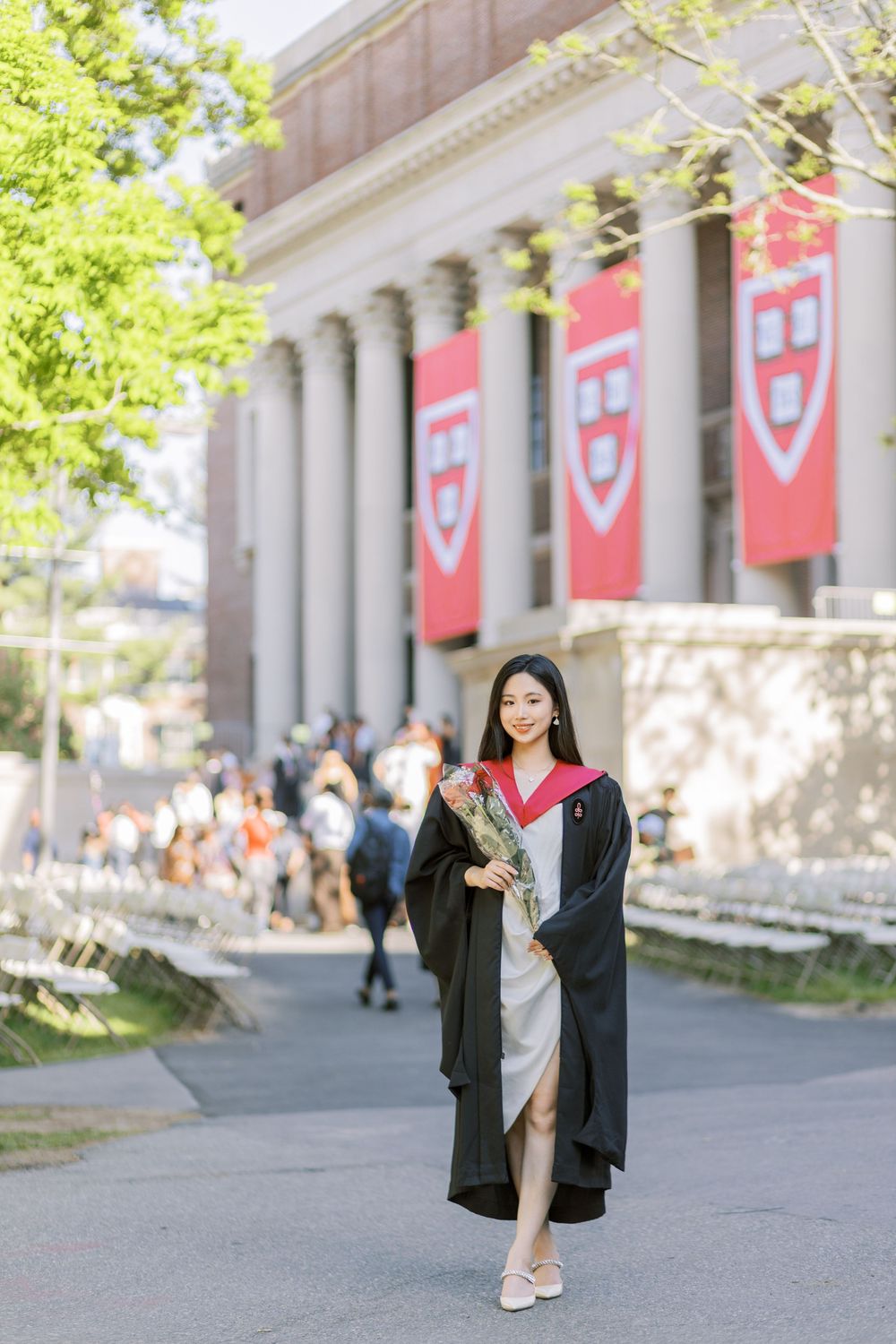 Harvard graduate in black gown holds roses outside Widener Library with red university banners in background.