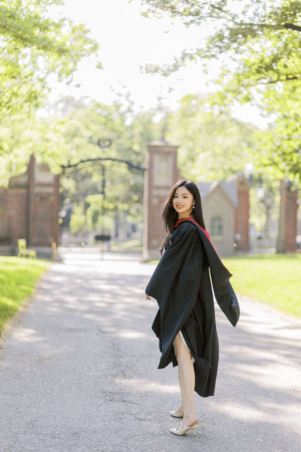Young woman in a Harvard graduation gown walks along tree-lined campus path near brick gate smiling.