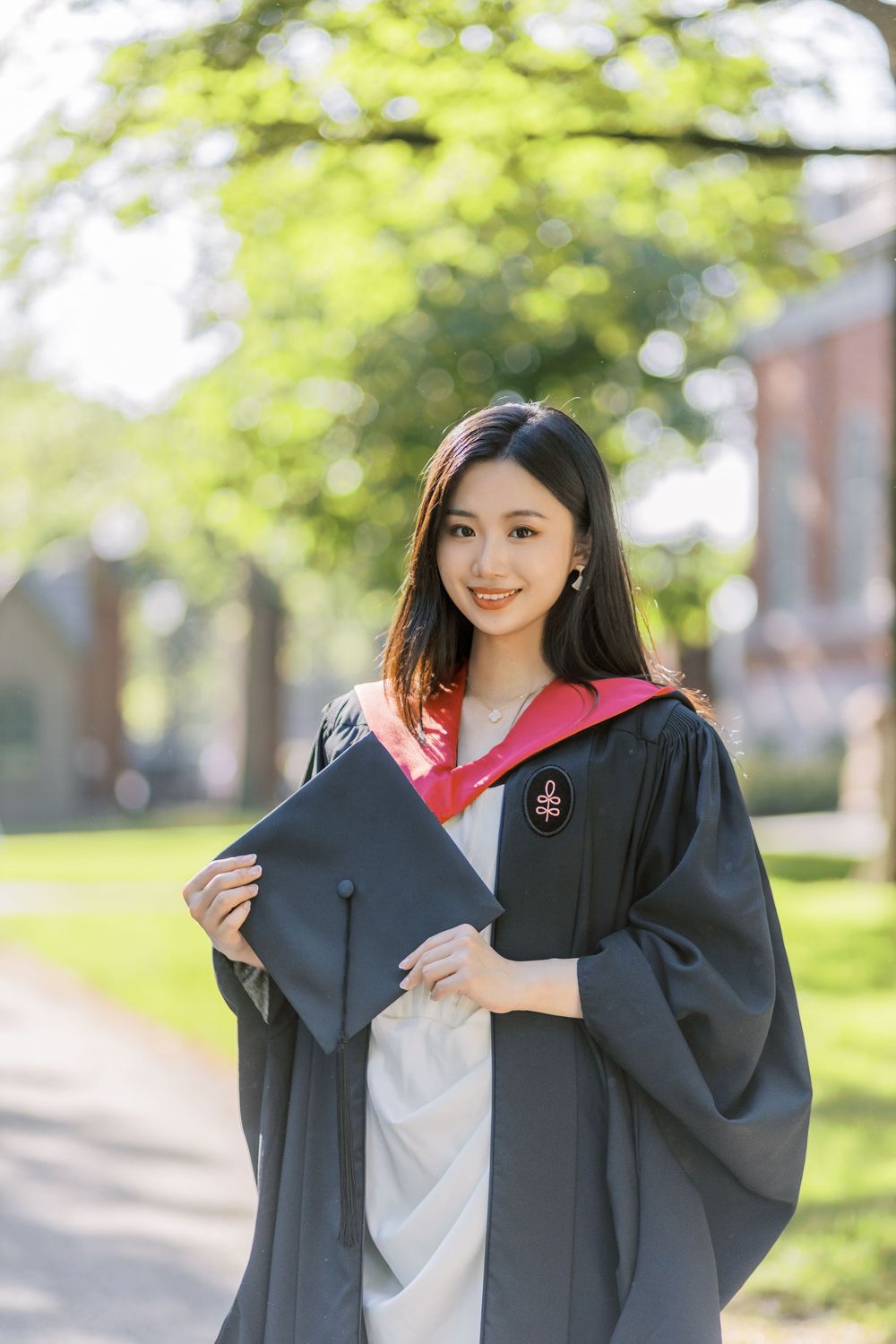 Smiling young woman in black Harvard graduation gown with pink hood holds cap on a tree-lined campus path.