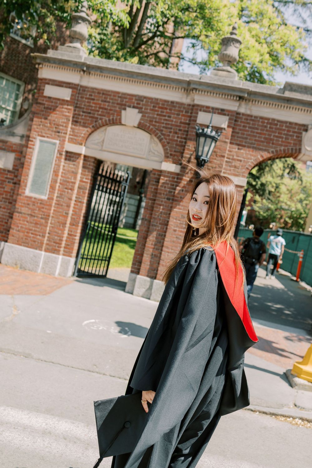 Young woman in a Harvard graduation gown with red sash poses outdoors near brick university gate on sunny day.