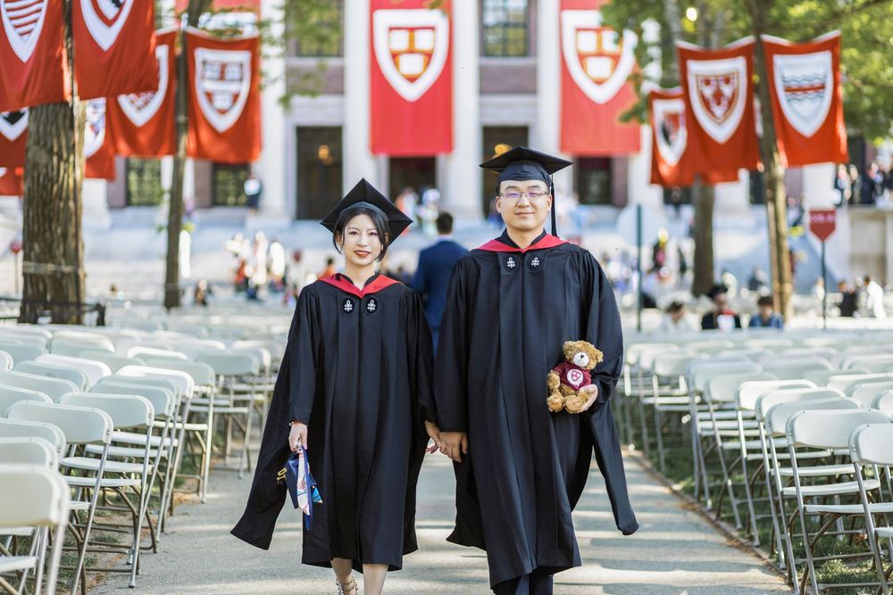 Two Harvard graduates in black caps and gowns walk together holding hands among red banners at commencement ceremony.