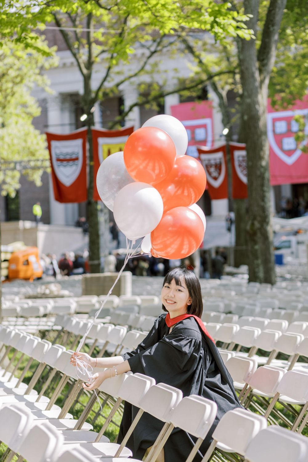 A Harvard graduate in black gown holds red and white balloons among white chairs at a Harvard commencement ceremony.