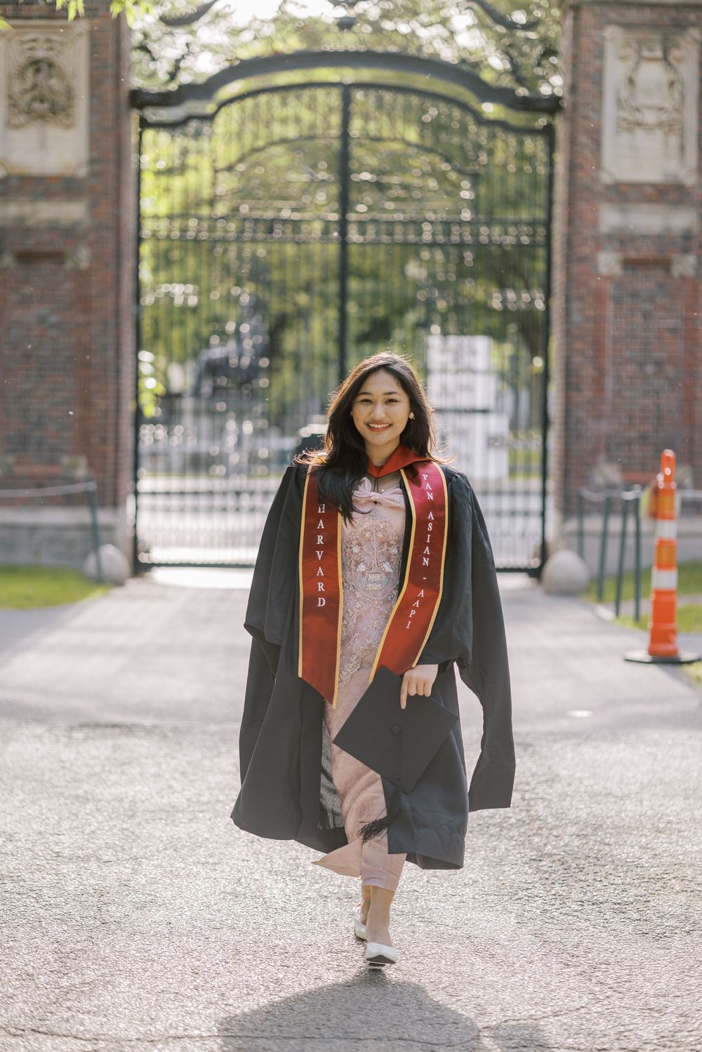 Young woman in black Harvard graduation gown with Harvard sash walks through ornate campus gates on a sunny day.