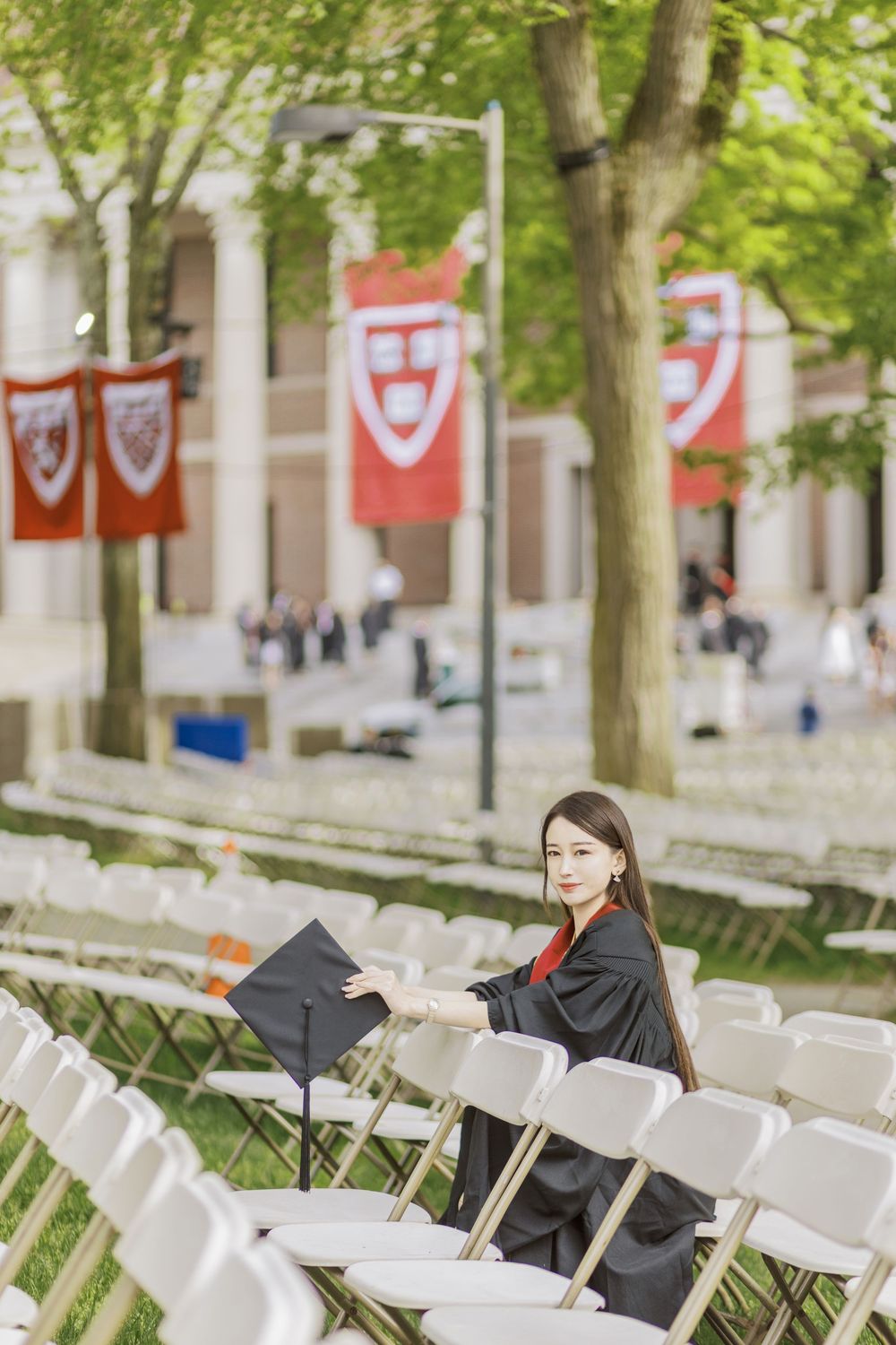 Harvard graduate in cap and gown poses among white chairs with crimson university banners in background.