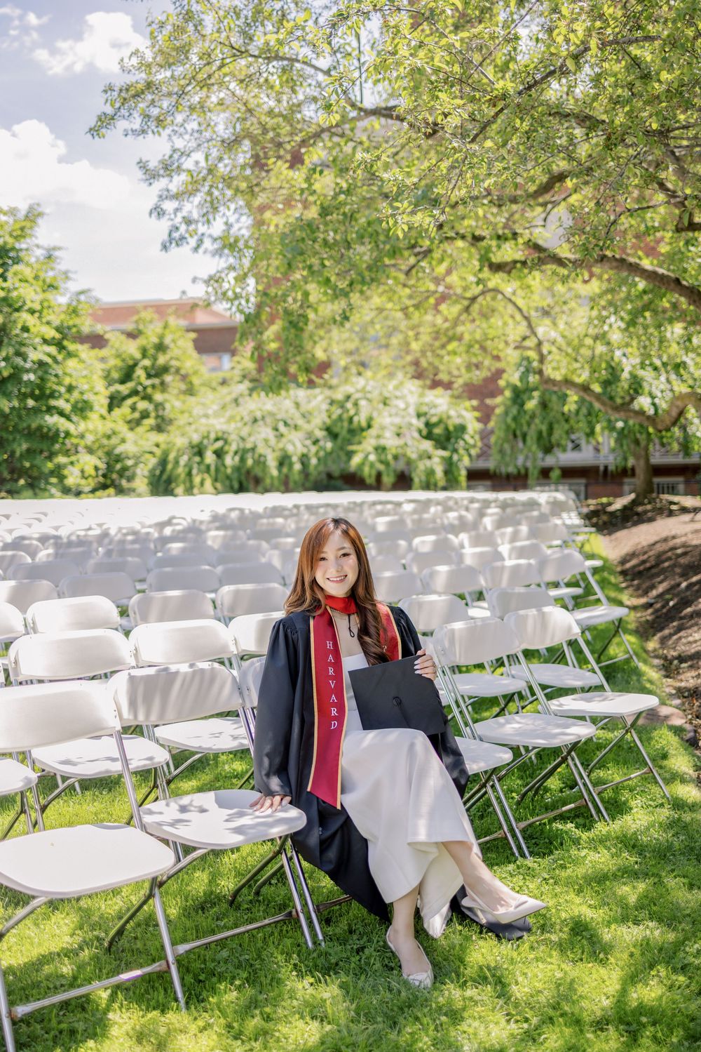 Smiling Harvard graduate in black cap and gown sits among white chairs outdoors holding diploma on sunny day.