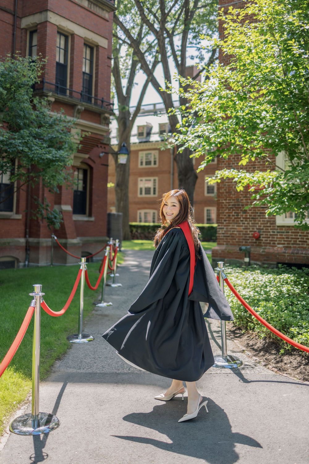 Young woman in a Harvard graduation gown with red sash poses on a sunlit campus pathway lined with red rope barriers.