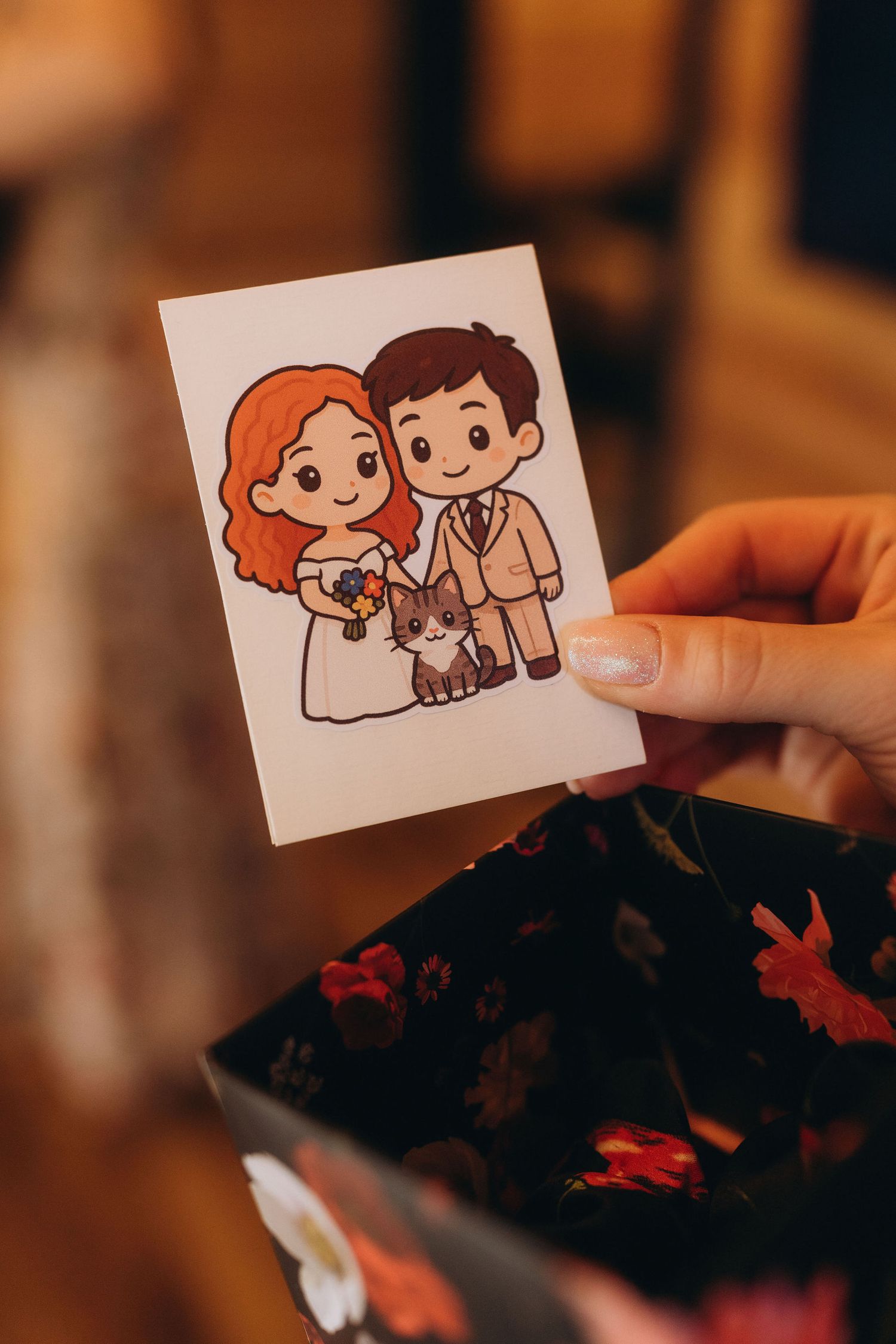 Hand holding a cute cartoon illustration of a bride and groom with a cat on a floral box.