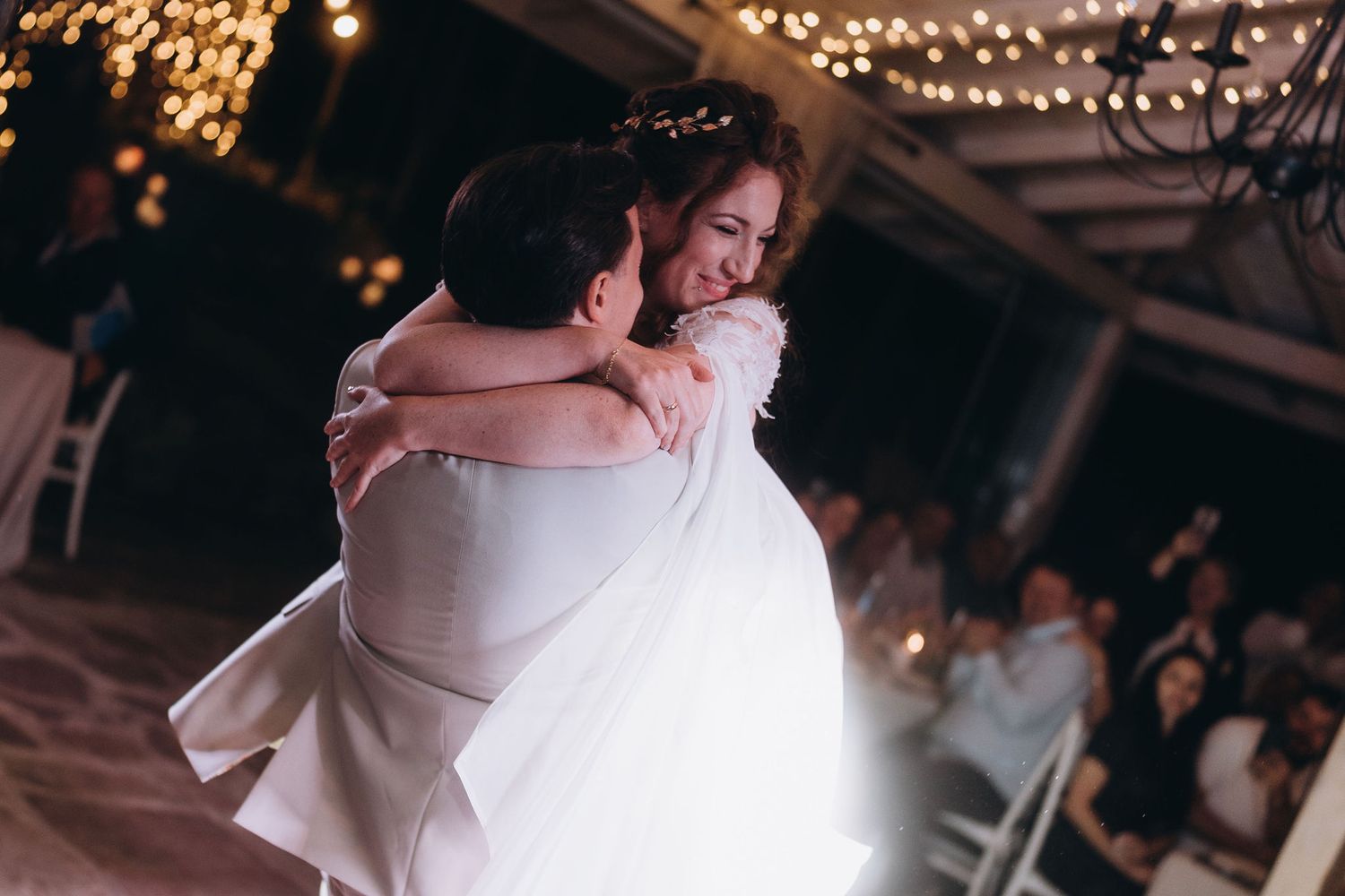 Groom lifts smiling bride in white wedding dress during joyful first dance at evening reception.