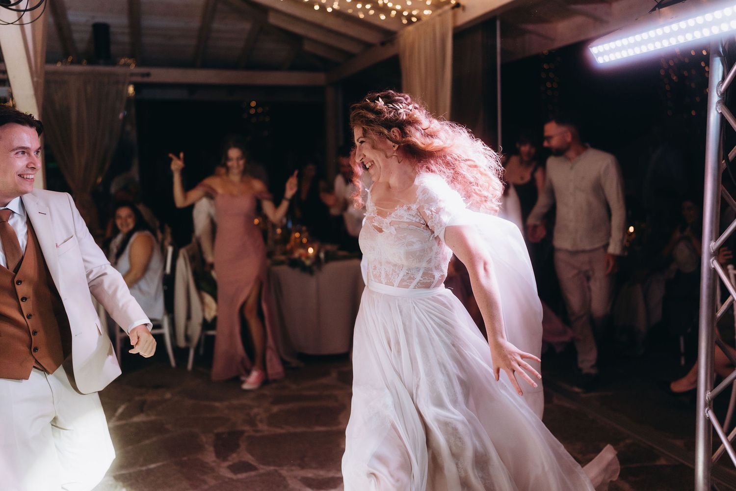 Bride in flowing white lace gown dancing joyfully with guests during lively outdoor wedding reception at night.