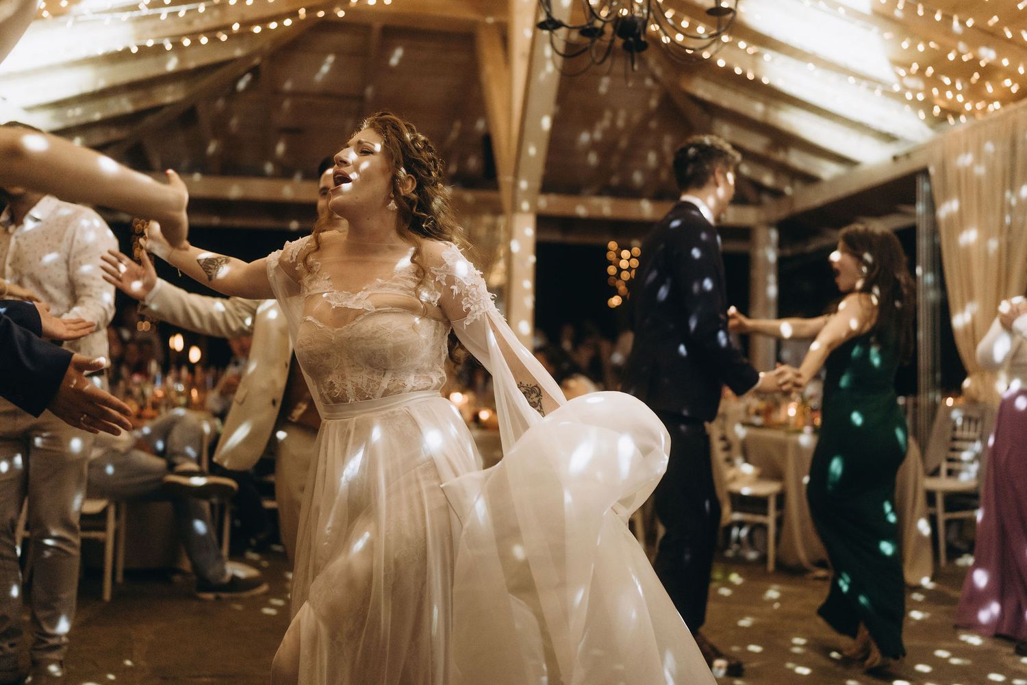 Bride twirls in lace wedding dress on dance floor surrounded by bubbles and fairy lights at indoor reception.