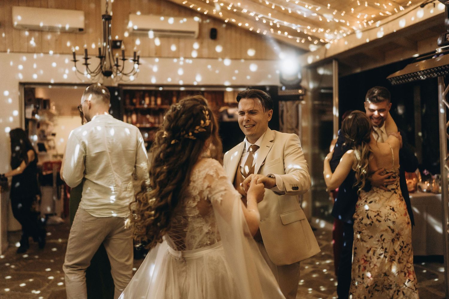 Bride and groom share first dance at elegant wedding reception with fairy lights and celebrating guests.