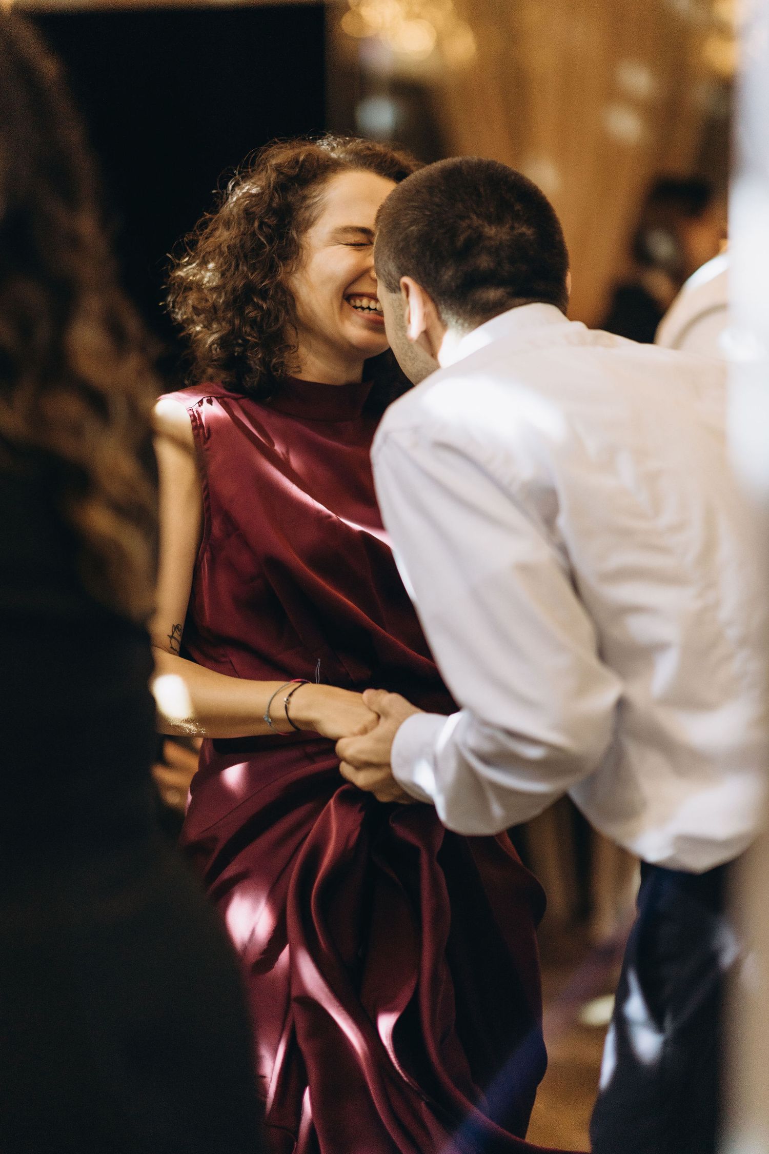 A joyful woman in a red satin dress laughs while dancing with a man in a white shirt at an elegant event.