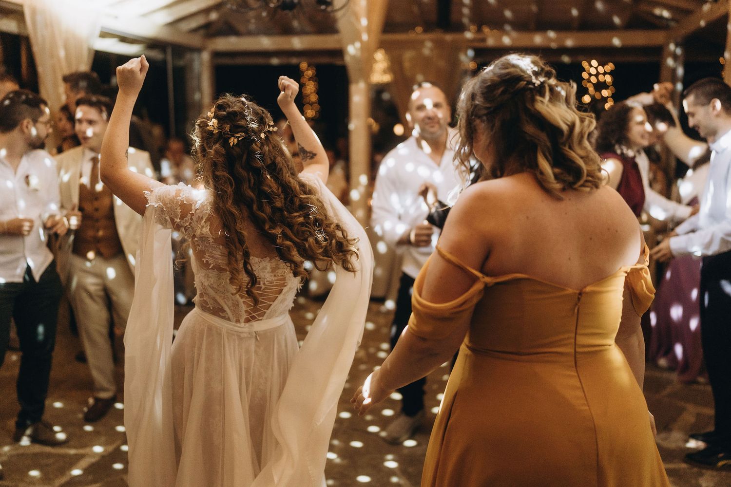 Bride in boho lace dress dances joyfully with guests under sparkling lights at wedding reception.
