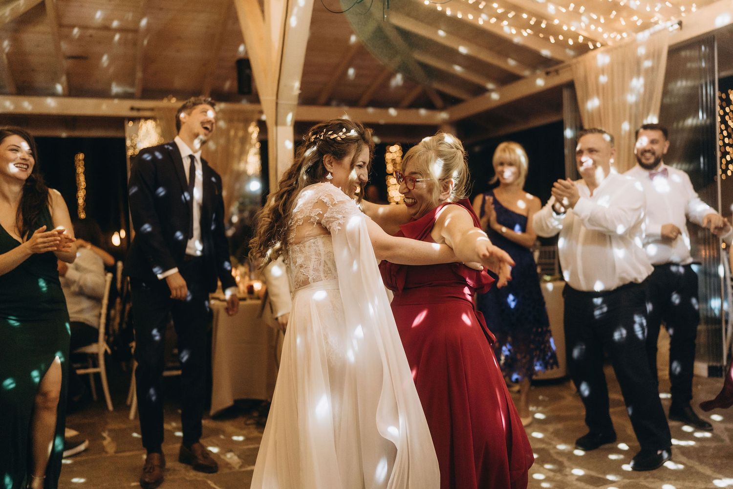 Bride dancing with woman in red dress at elegant wedding reception with fairy lights and bubbles.
