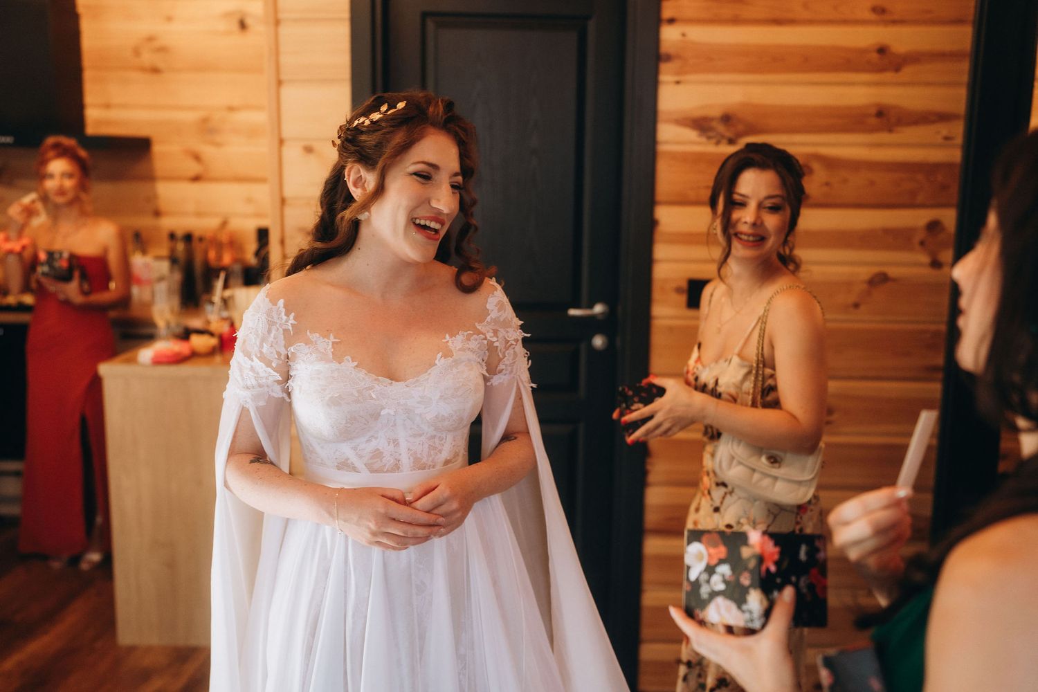 Bride in white wedding dress laughing with bridesmaids in a rustic wooden cabin getting-ready room.