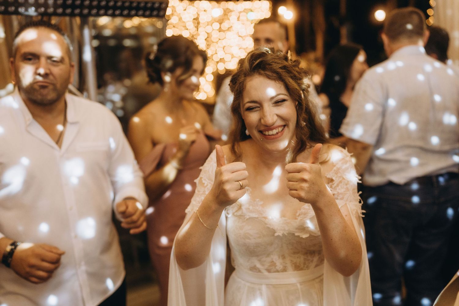 Bride in white dress giving thumbs up and smiling at wedding reception with bokeh lights.