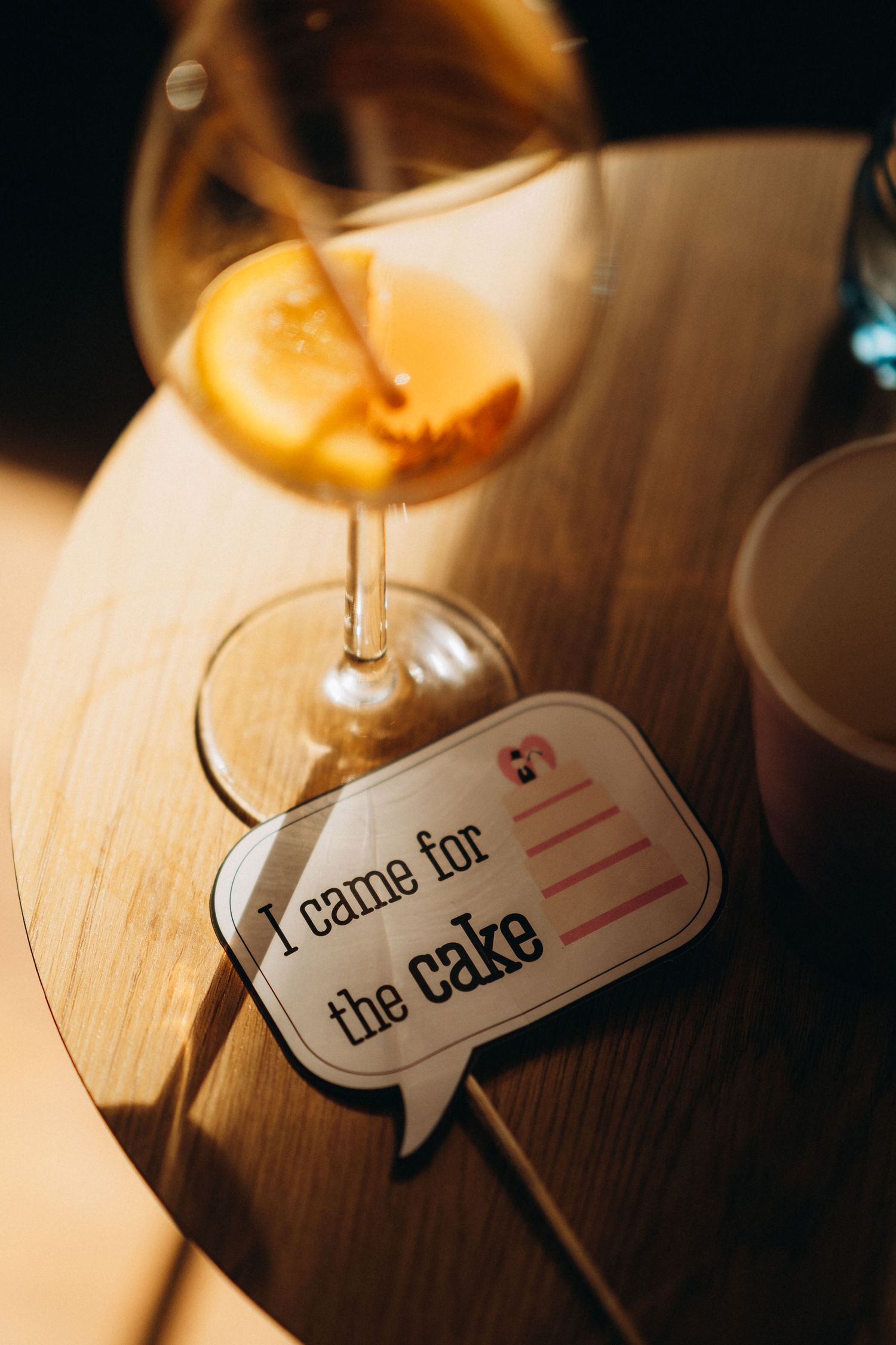 A speech bubble sign reading I came for the cake beside a cocktail with orange slice on a wooden table.