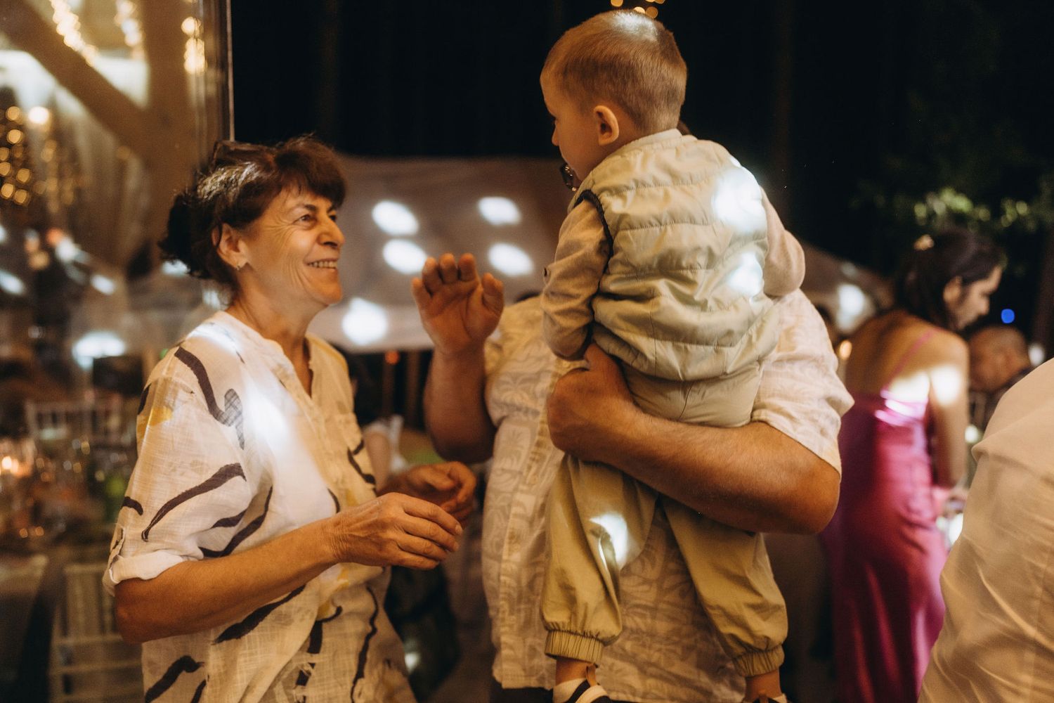 Elderly woman smiling and interacting with a baby held by an adult at a warmly lit wedding reception.