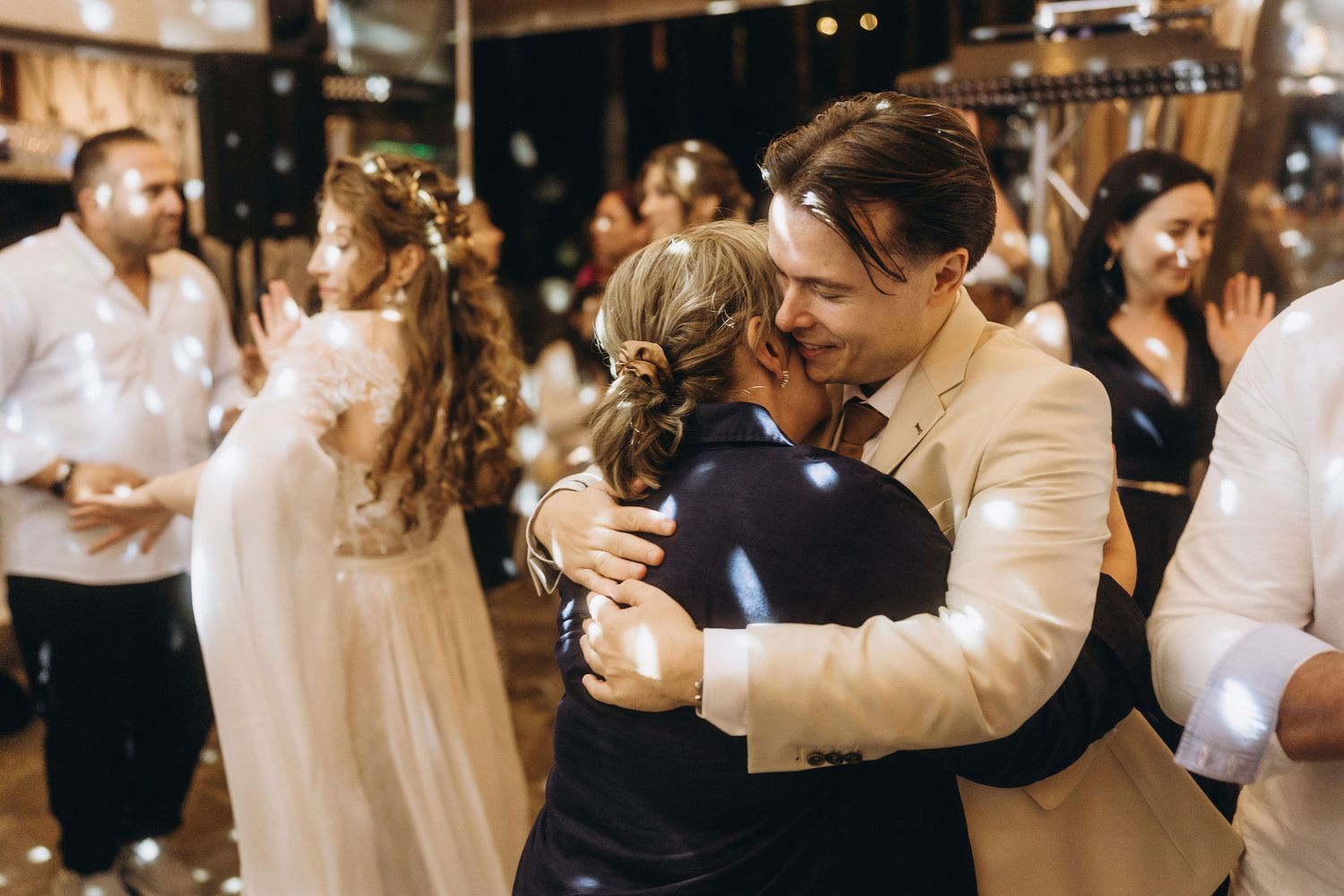 Groom in beige suit sharing warm embrace with guest on wedding reception dance floor.