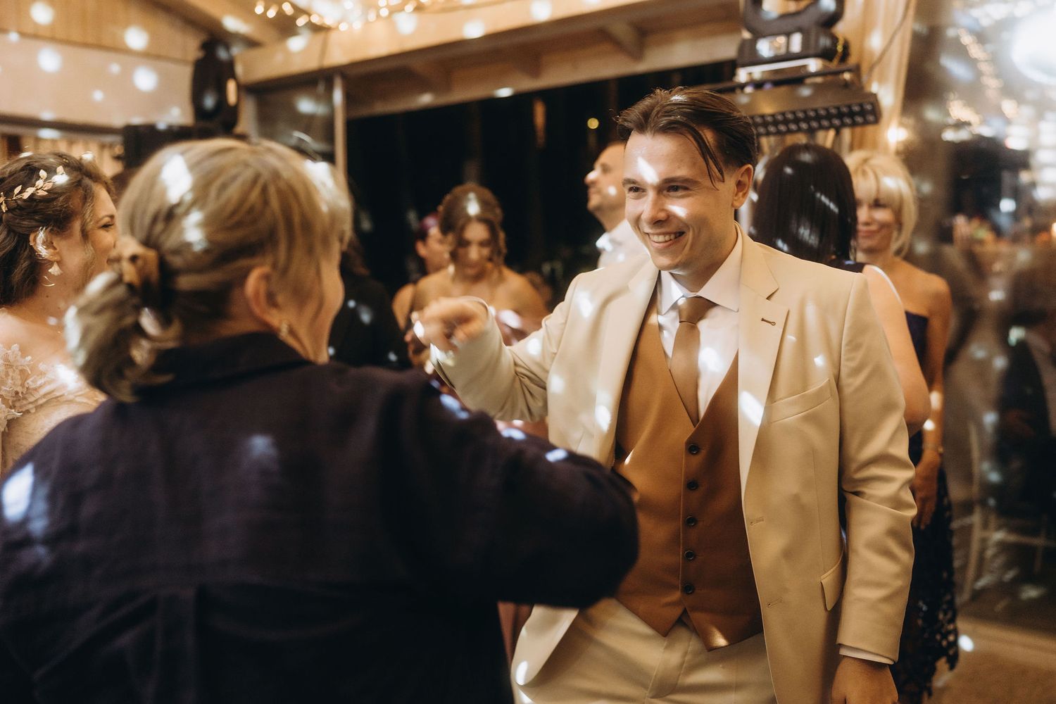 Smiling groom in tan suit with brown tie dancing with guest at wedding celebration.