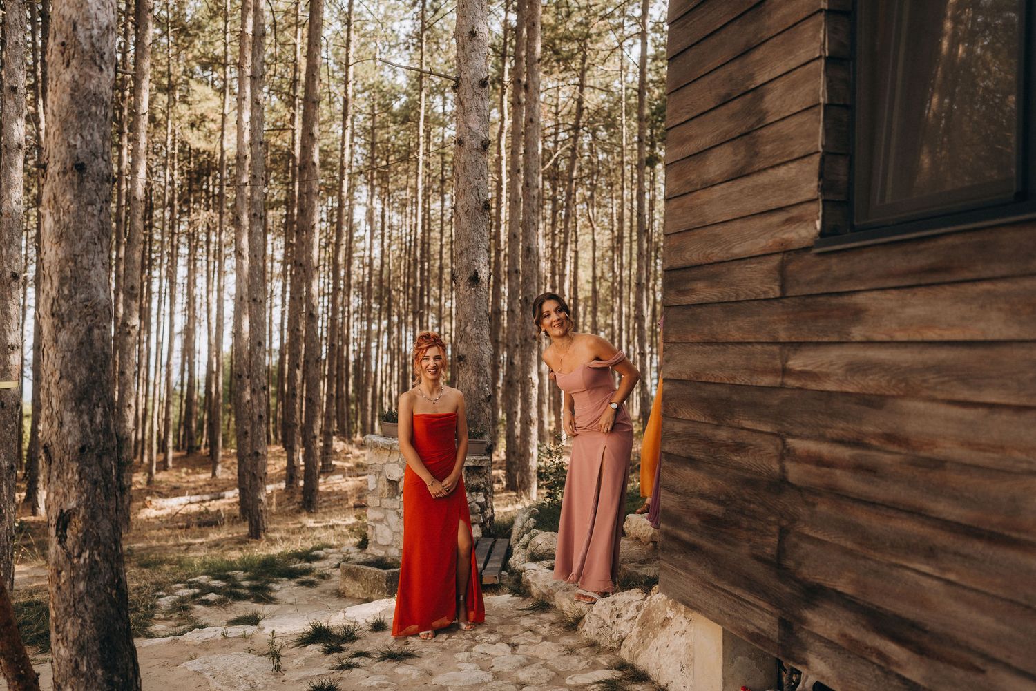 Two women in elegant red and pink gowns standing in a pine forest beside a rustic wooden cabin.