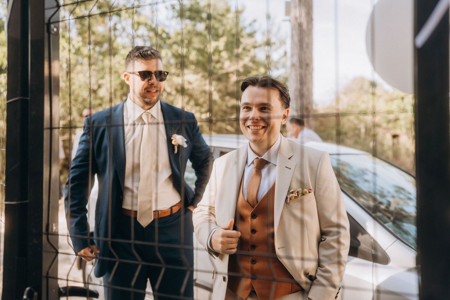 Two well-dressed grooms smile outdoors in wedding suits, one in navy blue and one in beige and brown.