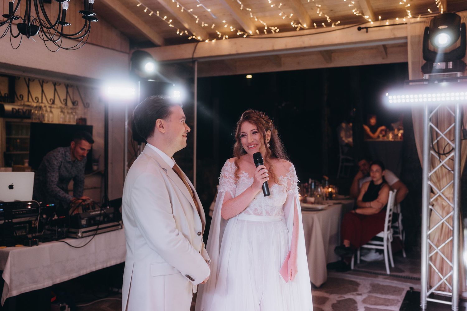 Bride speaking into microphone beside groom at elegant wedding reception with DJ and string lights.