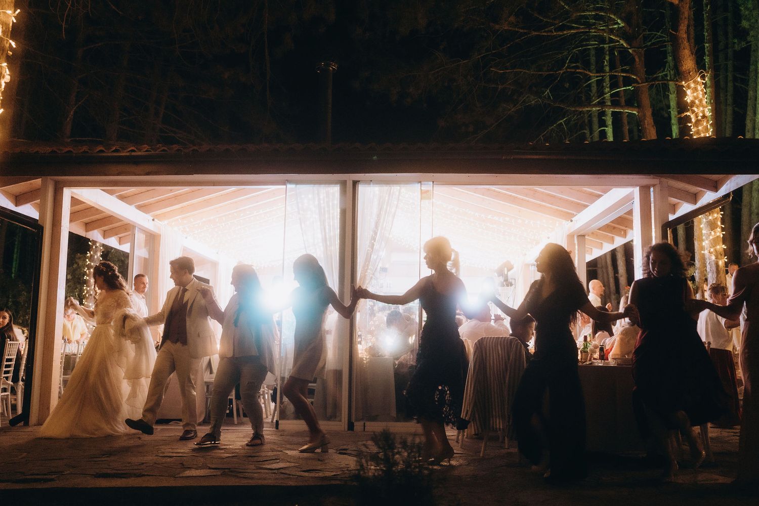 Silhouettes of wedding guests dancing and holding hands at an outdoor evening reception with lights.