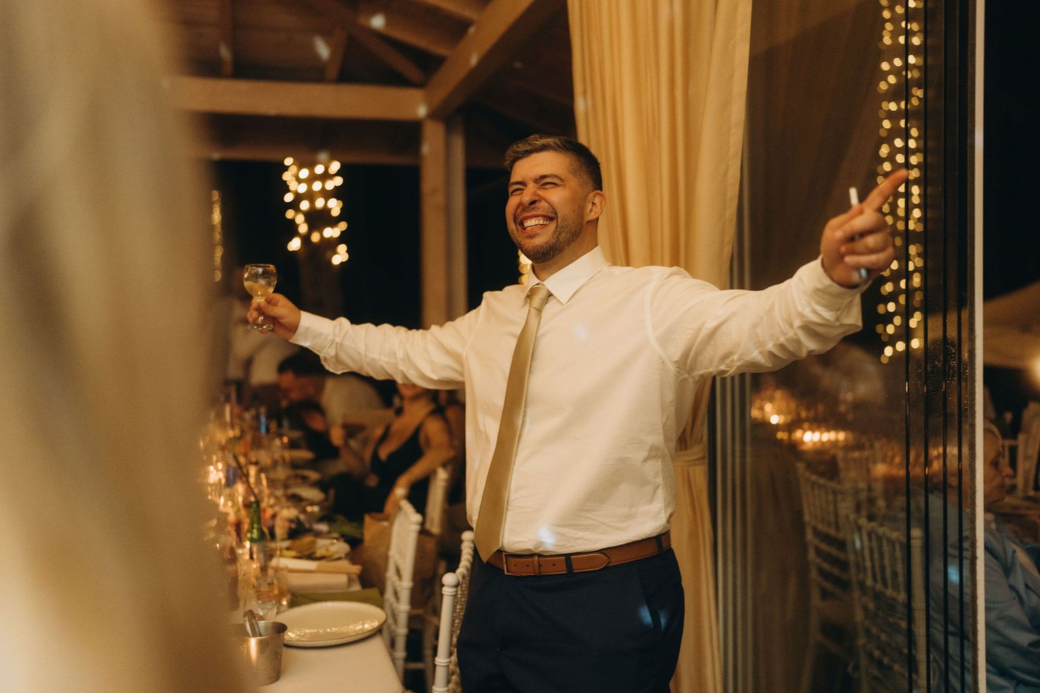 Smiling groomsman in white shirt and gold tie raises glass for toast at elegant wedding reception.