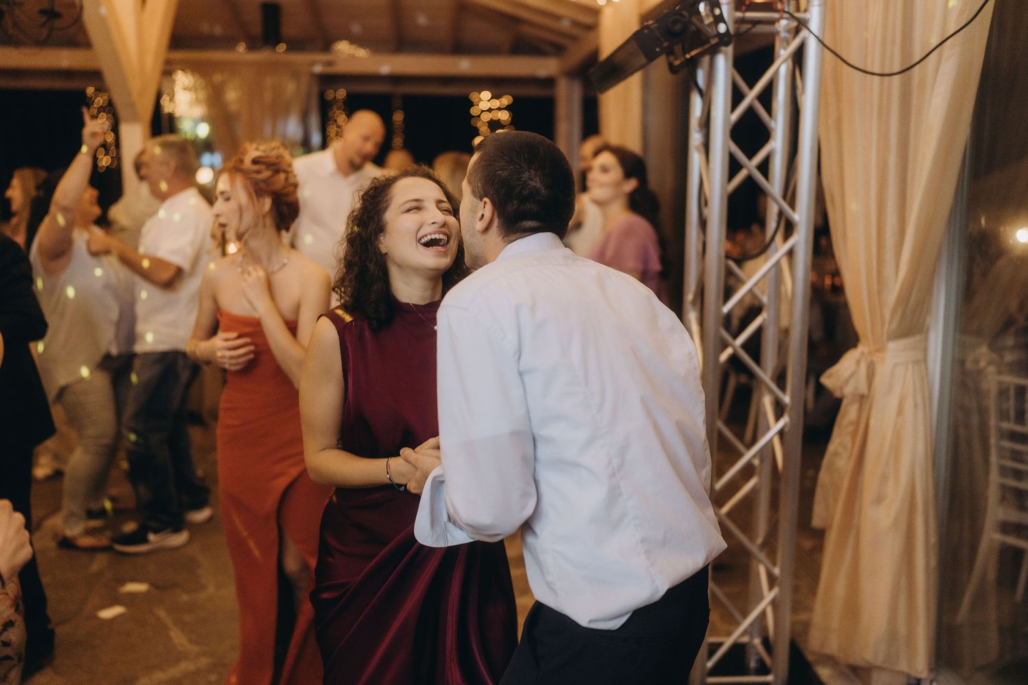 Laughing couple dances joyfully together at lively indoor wedding reception with festive lighting.