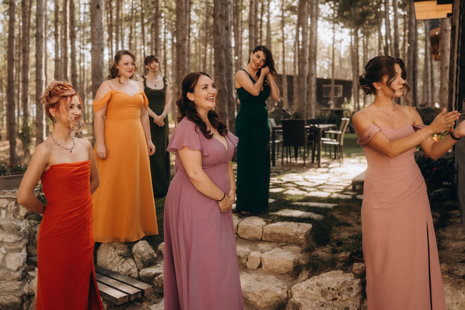 Bridesmaids in colorful mismatched dresses standing outdoors among pine trees at a rustic wedding venue.