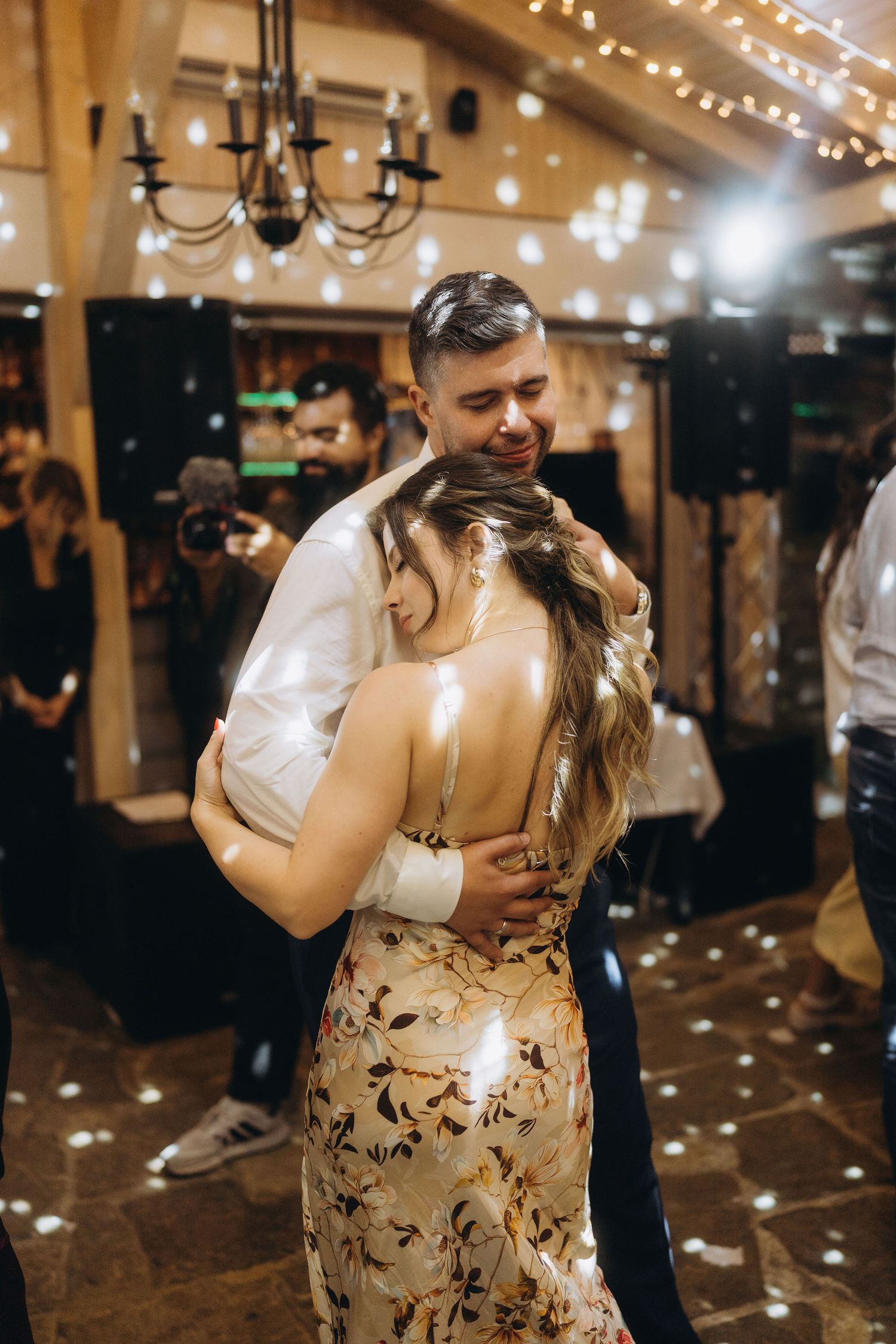 Couple sharing an intimate first dance at a wedding reception with warm string lights and chandelier in background.
