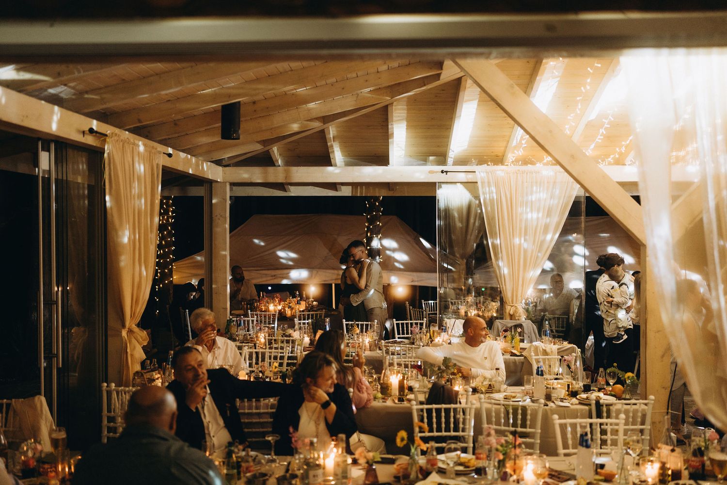 Warmly lit wedding reception venue with candles, fairy lights, draped fabric, and guests dining.