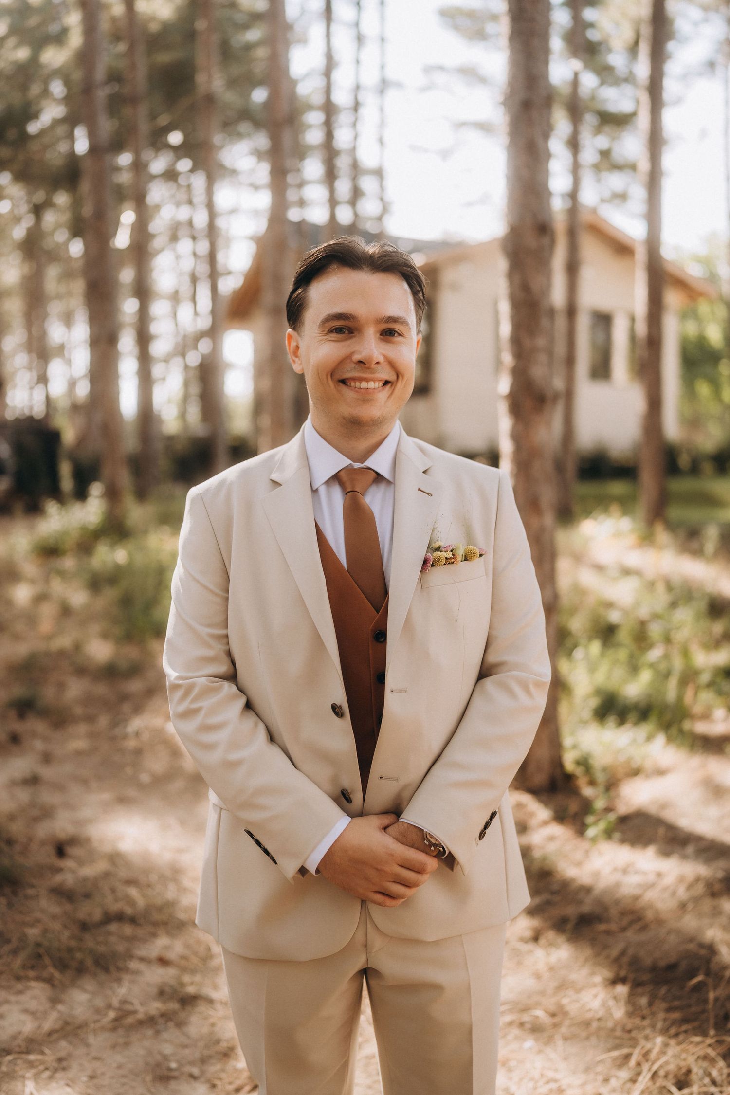 Smiling groom in beige suit with brown vest and tie stands outdoors among pine trees near a white building.