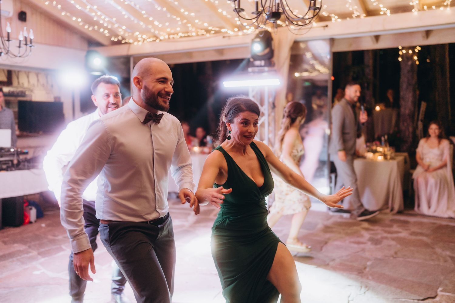 Couple dancing energetically at a wedding reception with string lights and guests watching in the background.