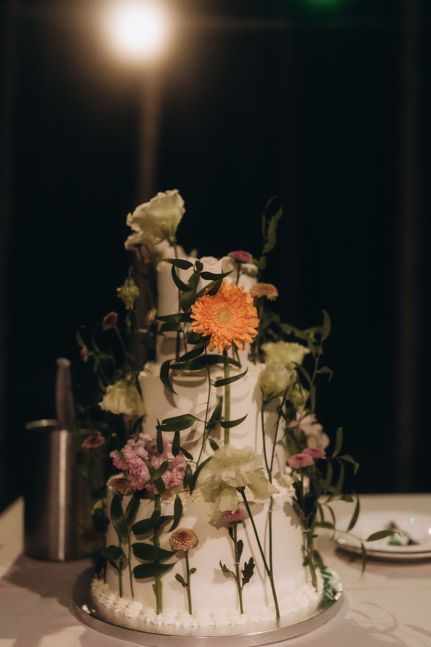 Elegant three-tier white wedding cake decorated with fresh colorful flowers and greenery on a dark background.