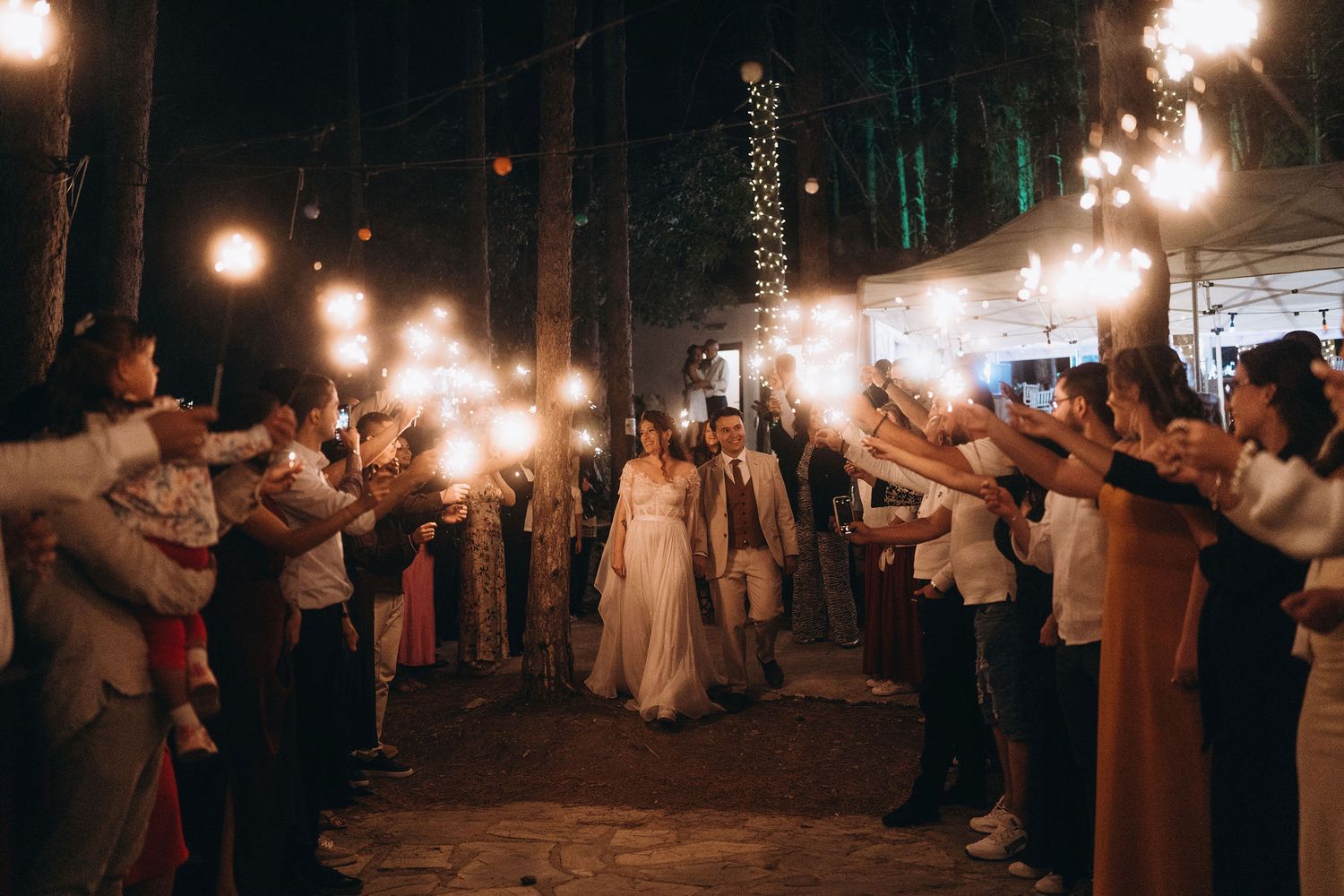 Bride and groom walk through a sparkler tunnel surrounded by cheering wedding guests at an outdoor night reception.