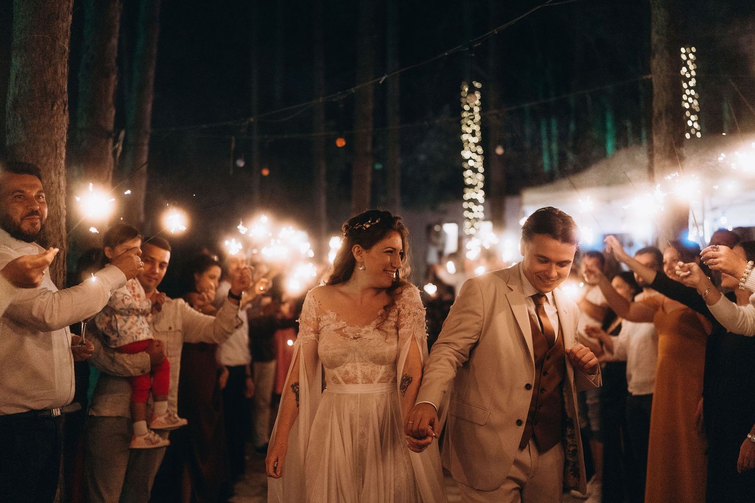Newlyweds holding hands walking through a glowing sparkler exit at their outdoor night wedding.