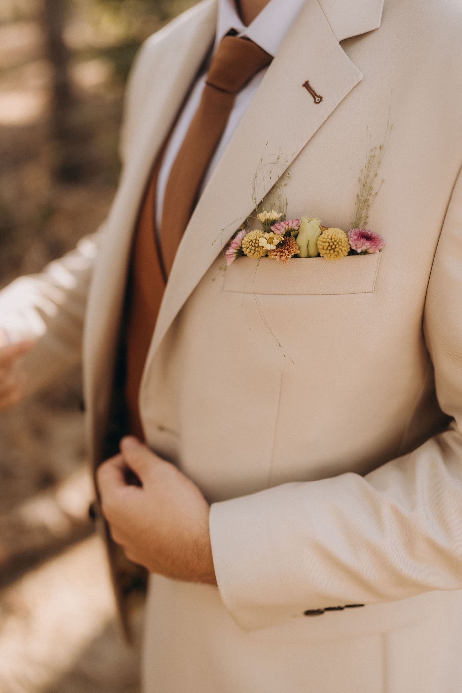 A groom in a beige suit with a brown tie and floral pocket square featuring small yellow and pink blooms.