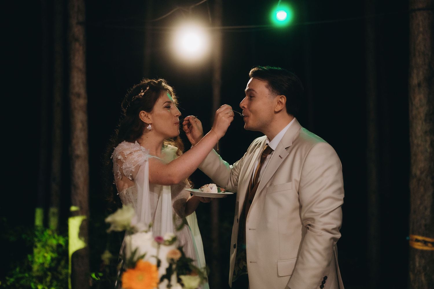 Bride feeding groom wedding cake during outdoor evening reception ceremony under warm lights.