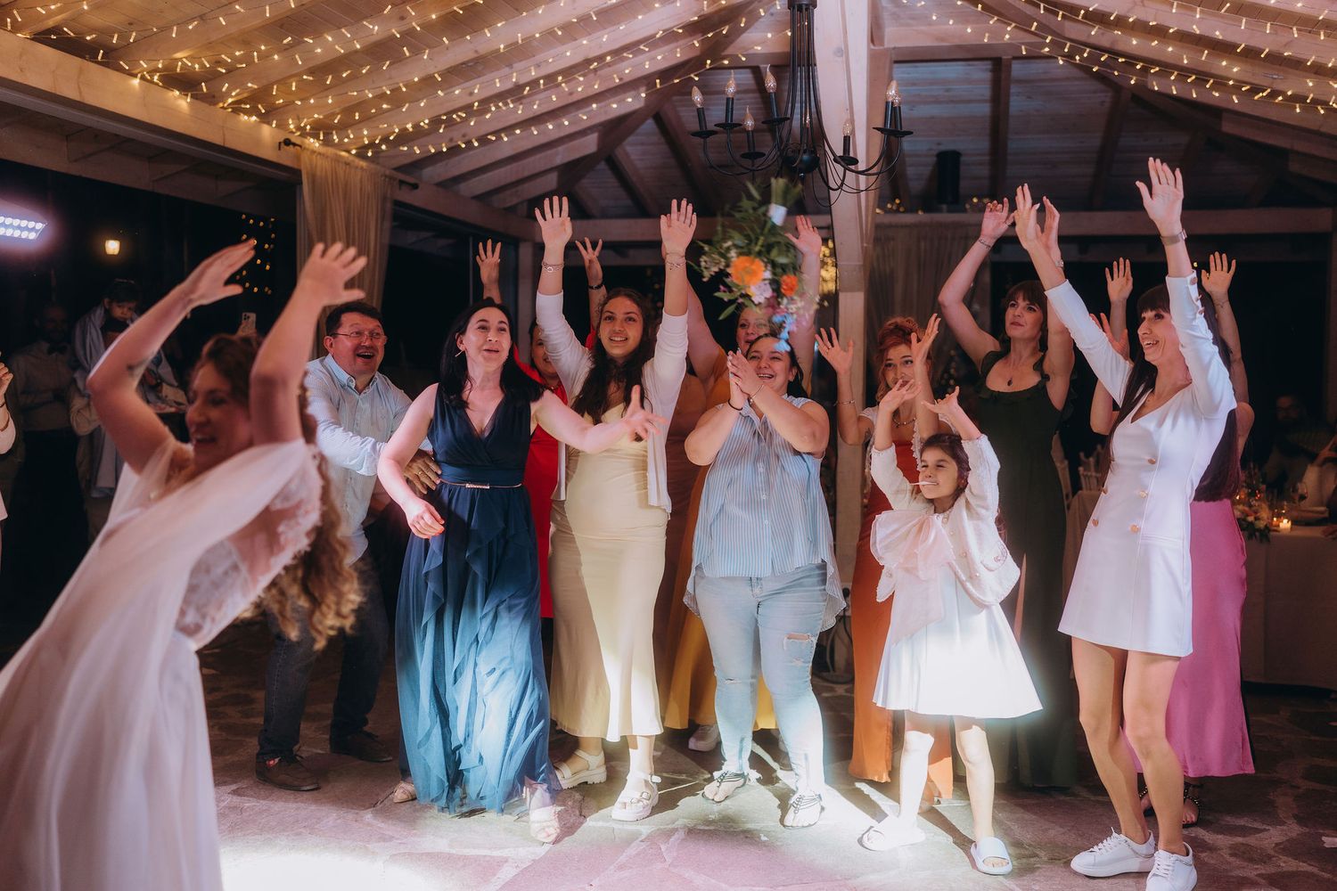Bride and wedding guests dancing with arms raised at a festive indoor reception with fairy lights.