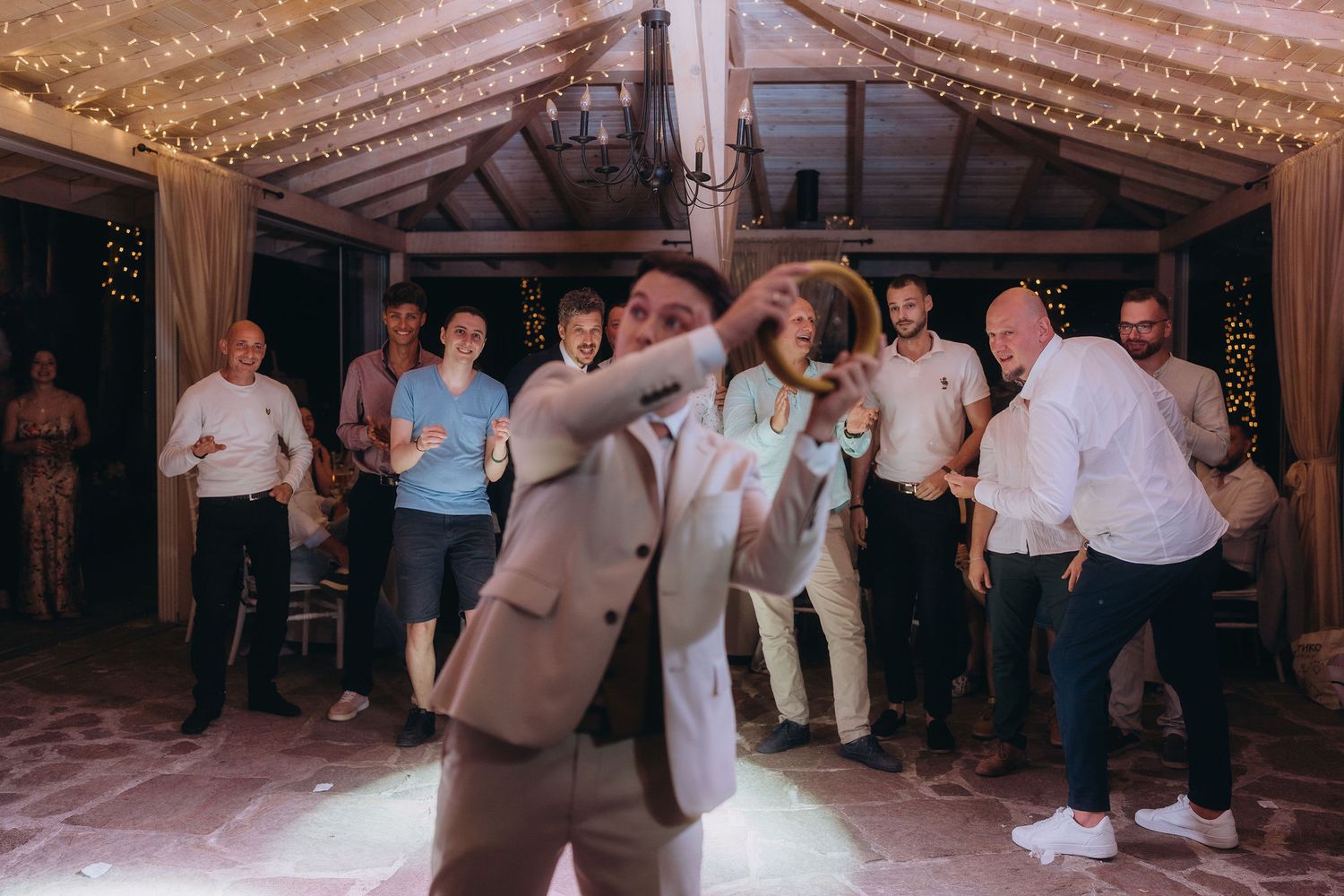 Groom dancing confidently at wedding reception while guests cheer in background under fairy lights.