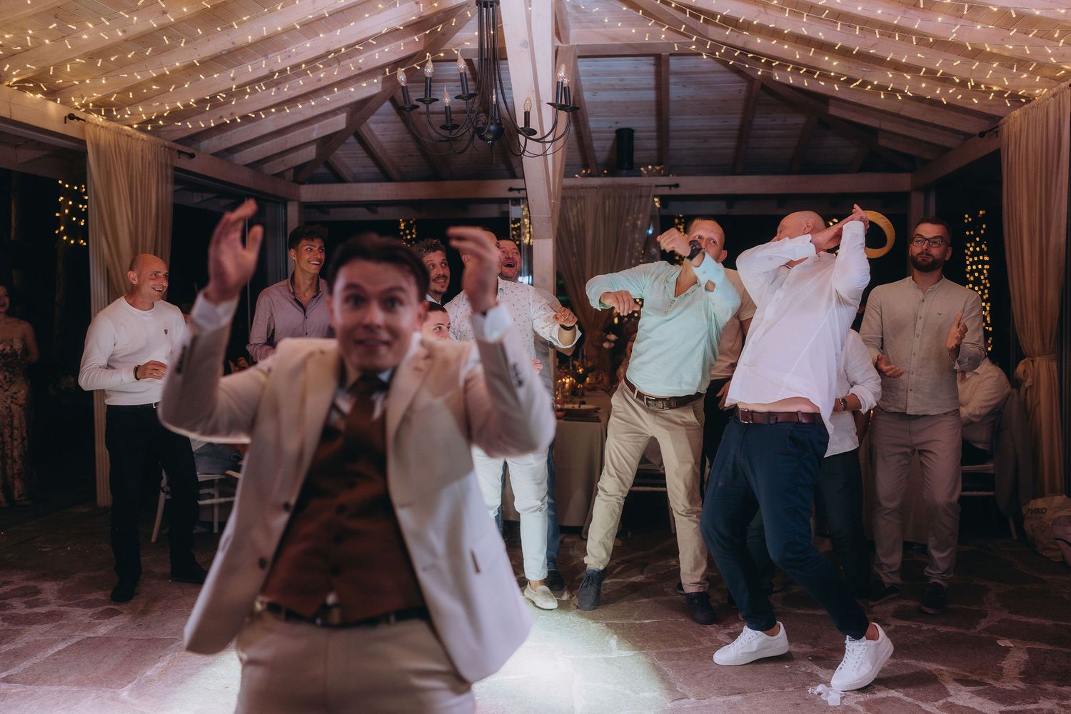 Group of men energetically dancing together at a wedding reception under fairy lights in a rustic venue.