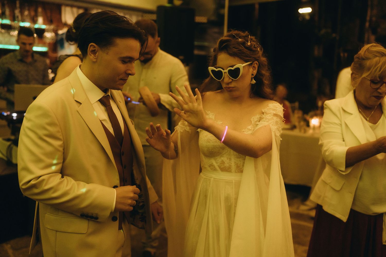 Couple dancing at wedding reception, bride in white gown with cat-eye sunglasses, groom in cream suit.