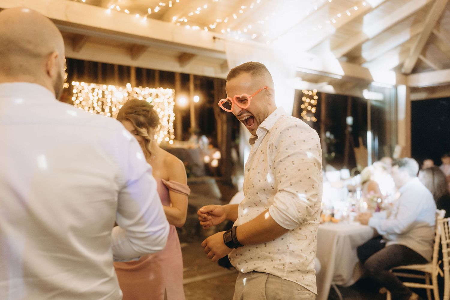 Guests dancing and laughing at a warm, festive wedding reception with string lights glowing in the background.